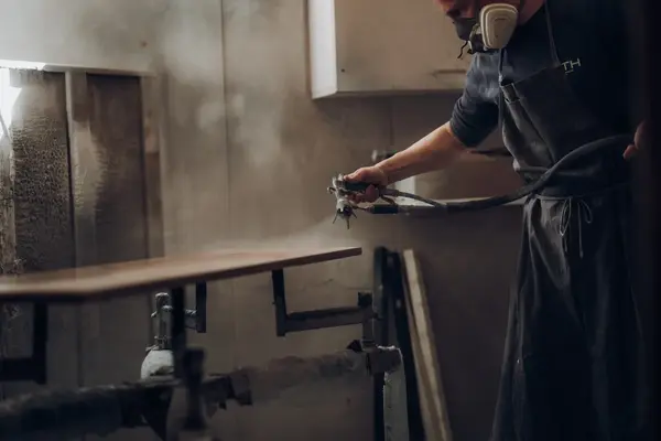 Maler sprüht Lack auf Holzplatte in Werkstatt, Atemschutz beim Lackieren