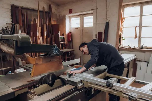 Tischler in Werkstatt sägt Holz an Formatkreissäge, Holzplatten im Hintergrund