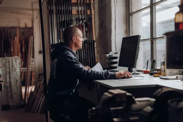 Schreiner im Holzwerkstatt-Büro arbeitet am PC am Fenster, Holzstangen im Hintergrund